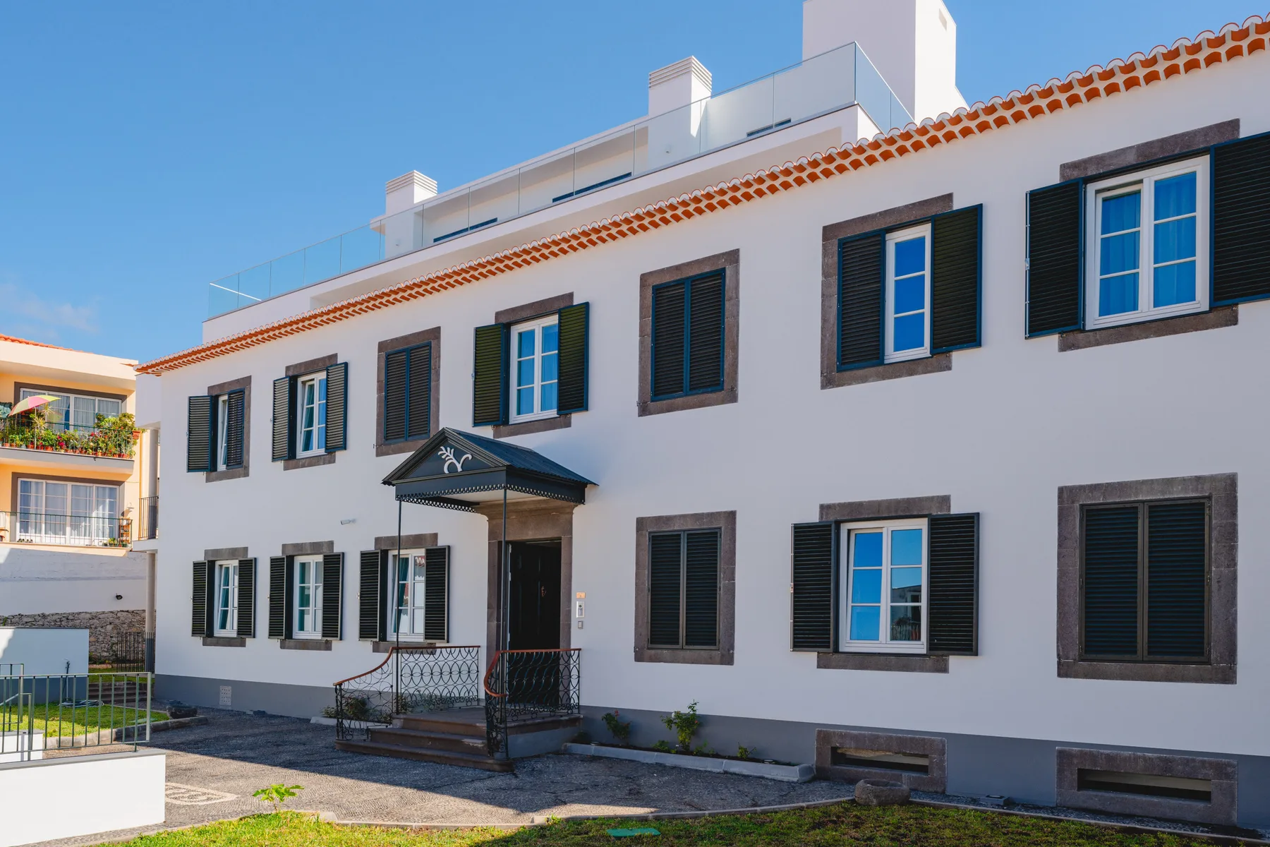 Edificio blanco de varias plantas con contraventanas oscuras y una entrada cubierta.