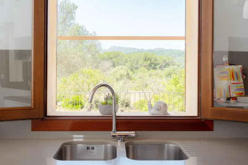 A kitchen sink sits beneath a large window overlooking green hills.