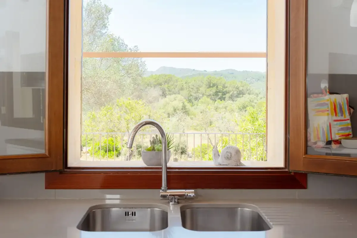 A kitchen sink sits beneath a window overlooking green hills.