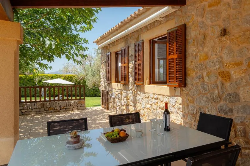 A shaded outdoor dining area beside a stone house, with a table and wooden shutters.