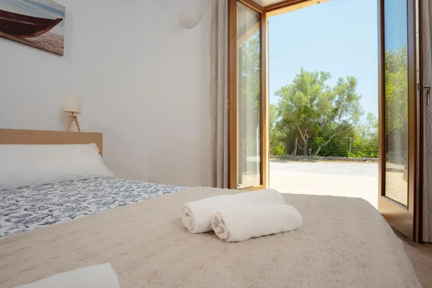 A bedroom with a bed and folded towels beside open glass doors overlooking greenery.
