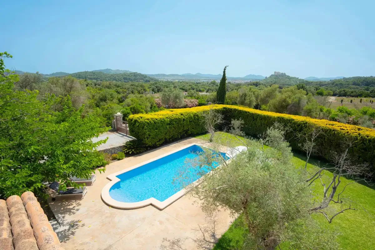 An outdoor swimming pool sits beside a hedge, with wooded hills in the distance.