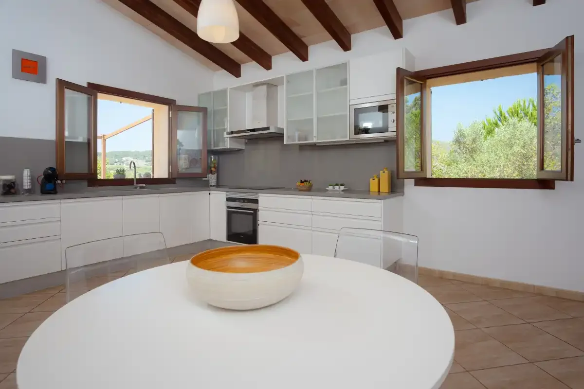 A bright kitchen with white cabinets, a central table, and open windows.