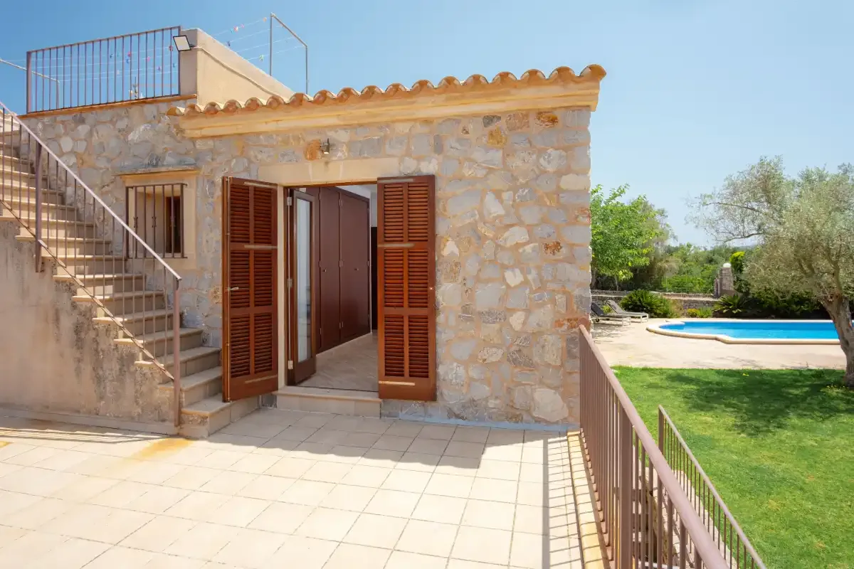 A stone building entrance opens onto a tiled terrace beside a swimming pool.