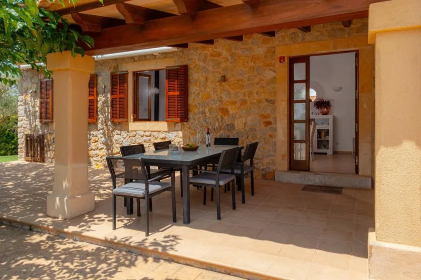 A shaded patio has a dining table and seating beside a stone exterior wall.