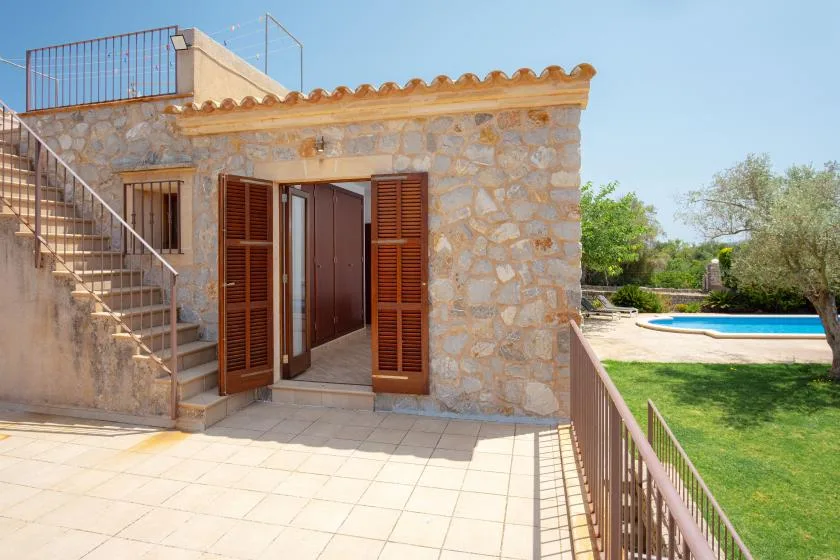 A stone building opens onto a tiled terrace overlooking a swimming pool and garden.