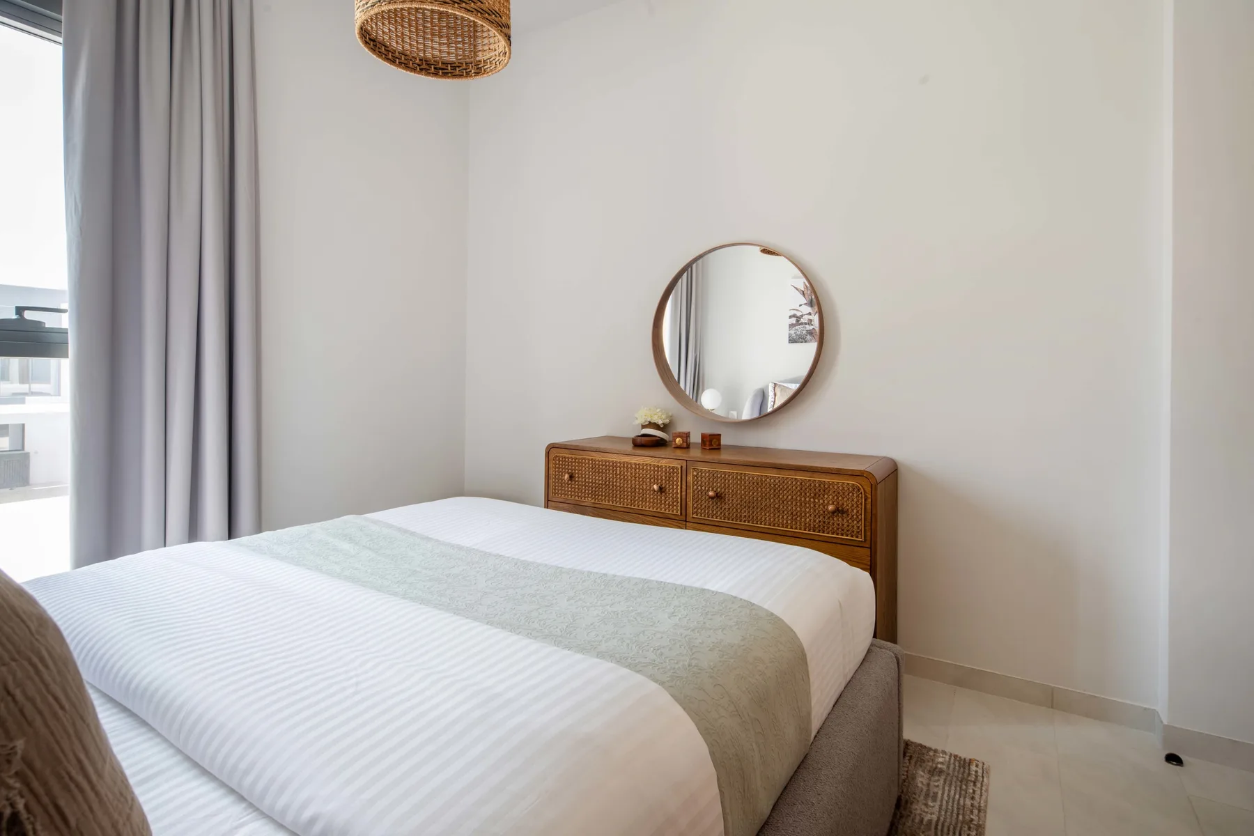 Un dormitorio con una cama bien tendida y una cómoda de madera bajo un espejo redondo.