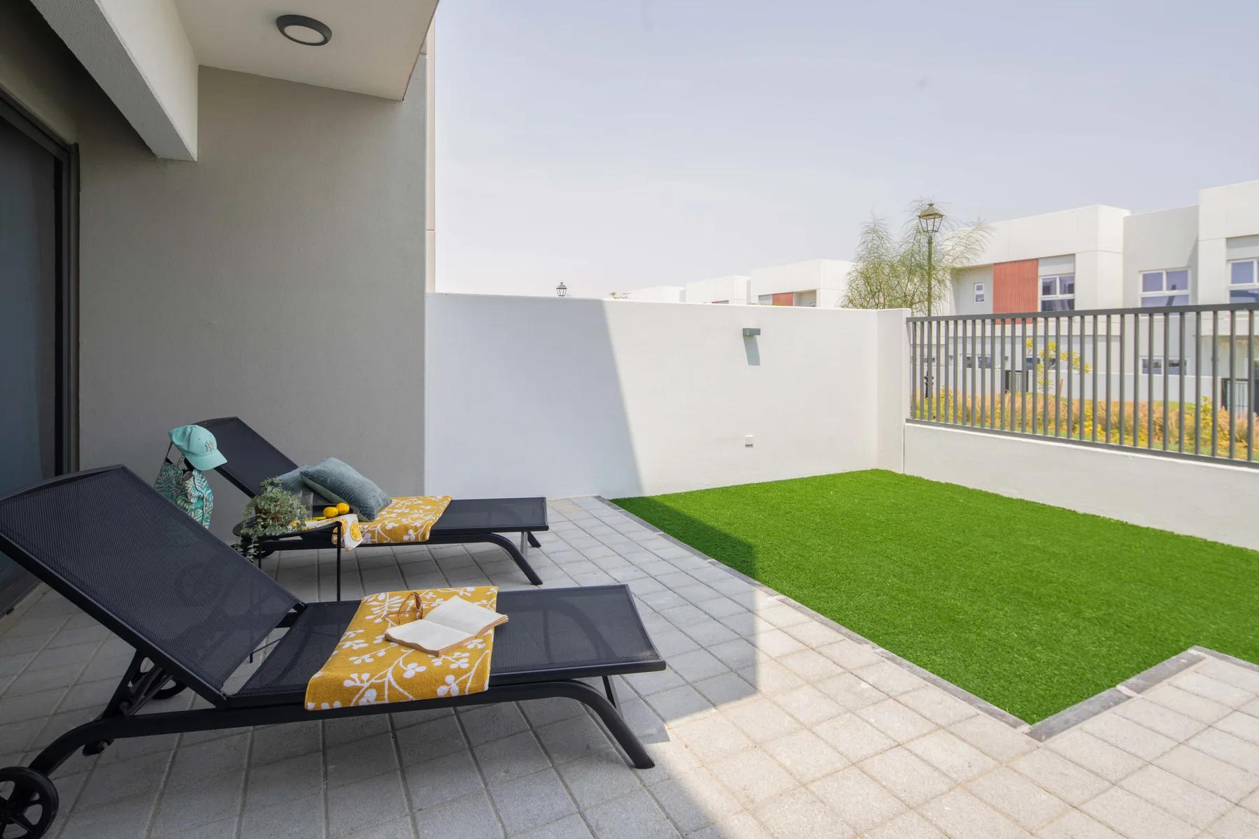 Un patio exterior embaldosado tiene asientos reclinables junto a una zona rectangular de césped.
