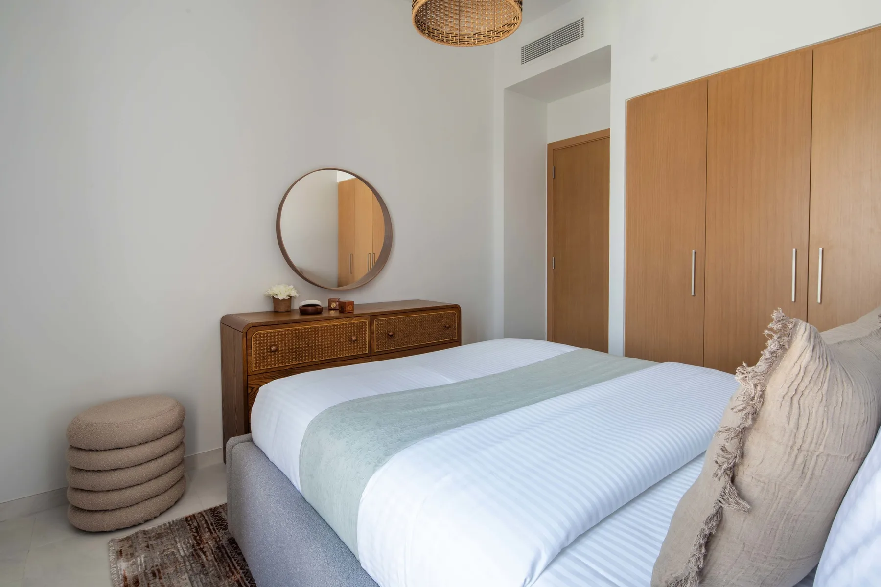 Dormitorio con una cama bien arreglada, armario de madera y espejo redondo en la pared.