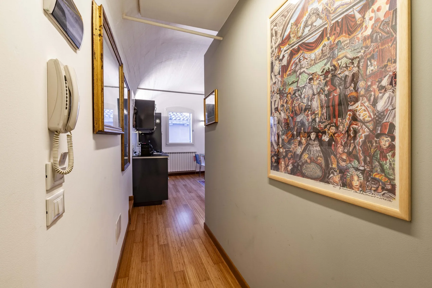 A narrow hallway with wood flooring and framed artwork along the walls.