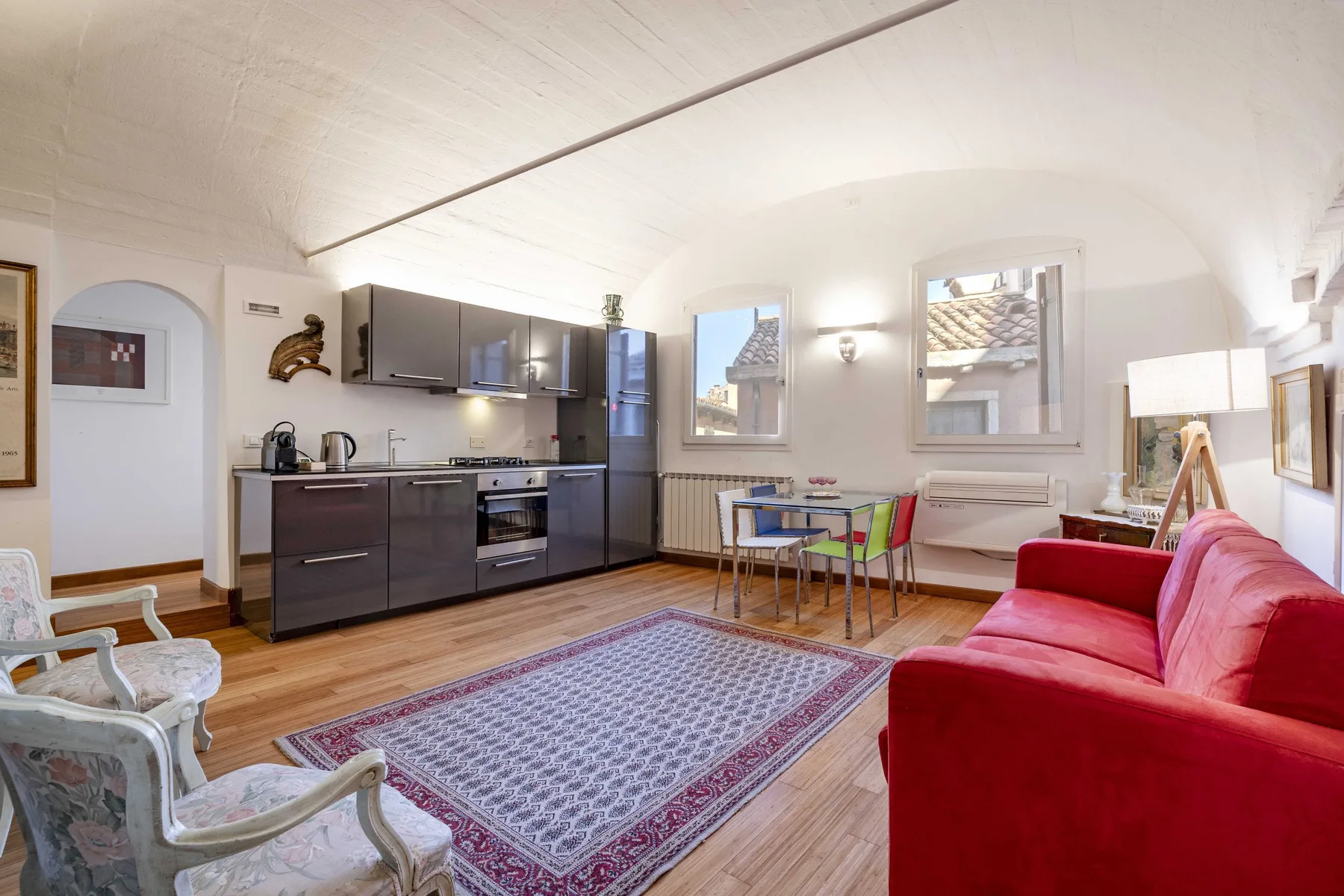 A bright living room with a kitchen area, a red sofa, and a patterned rug.