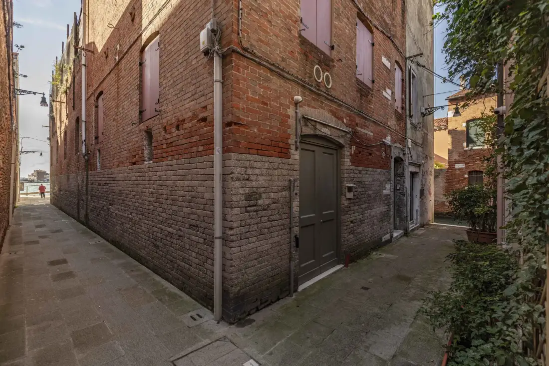 A narrow paved alley runs beside a brick building with a dark entrance door.