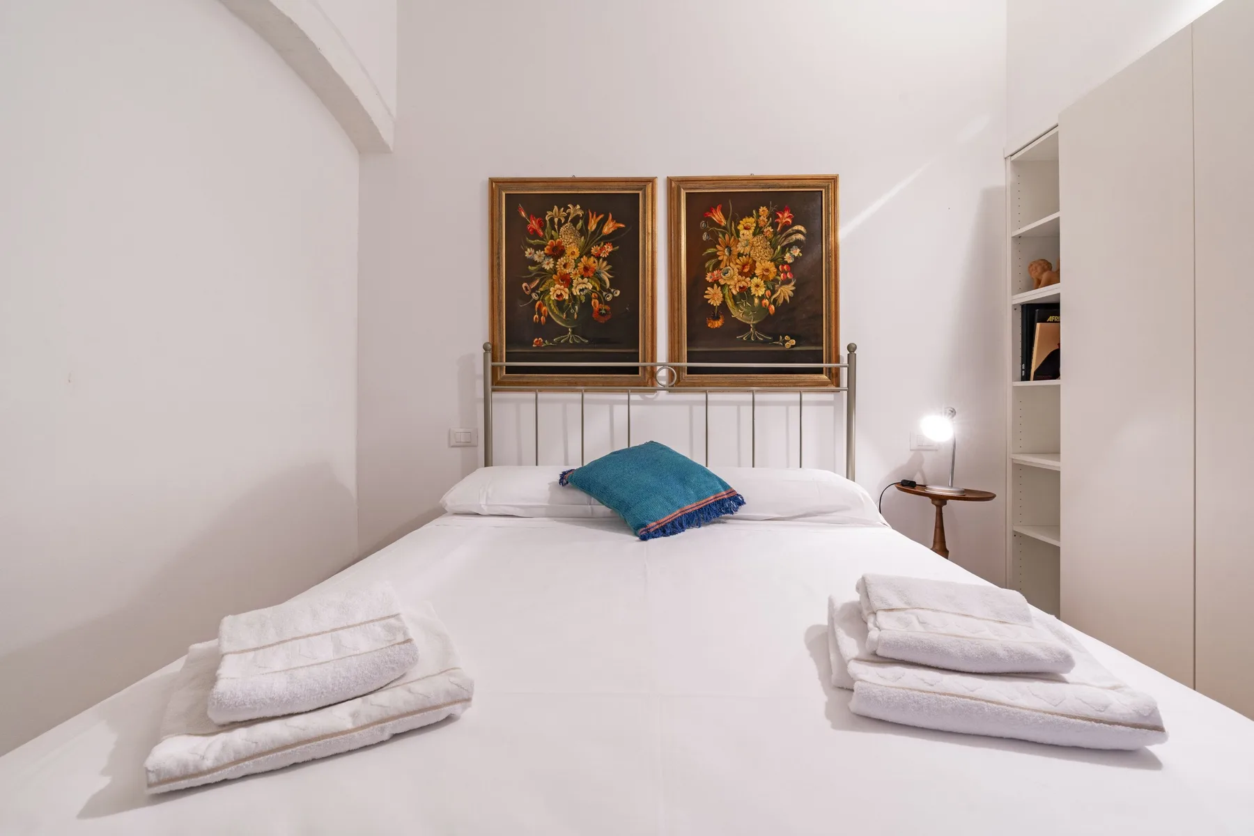 Un dormitorio con una cama blanca, un cojín turquesa y dos cuadros florales sobre el cabecero.