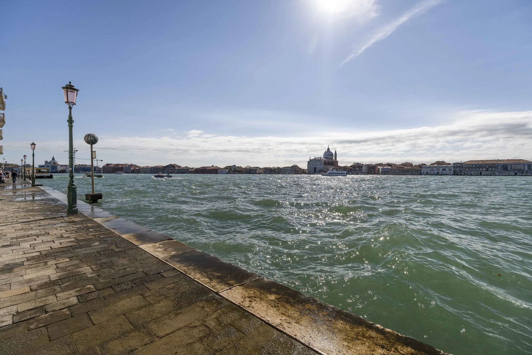 A waterfront promenade borders choppy water beneath a bright, partly cloudy sky.