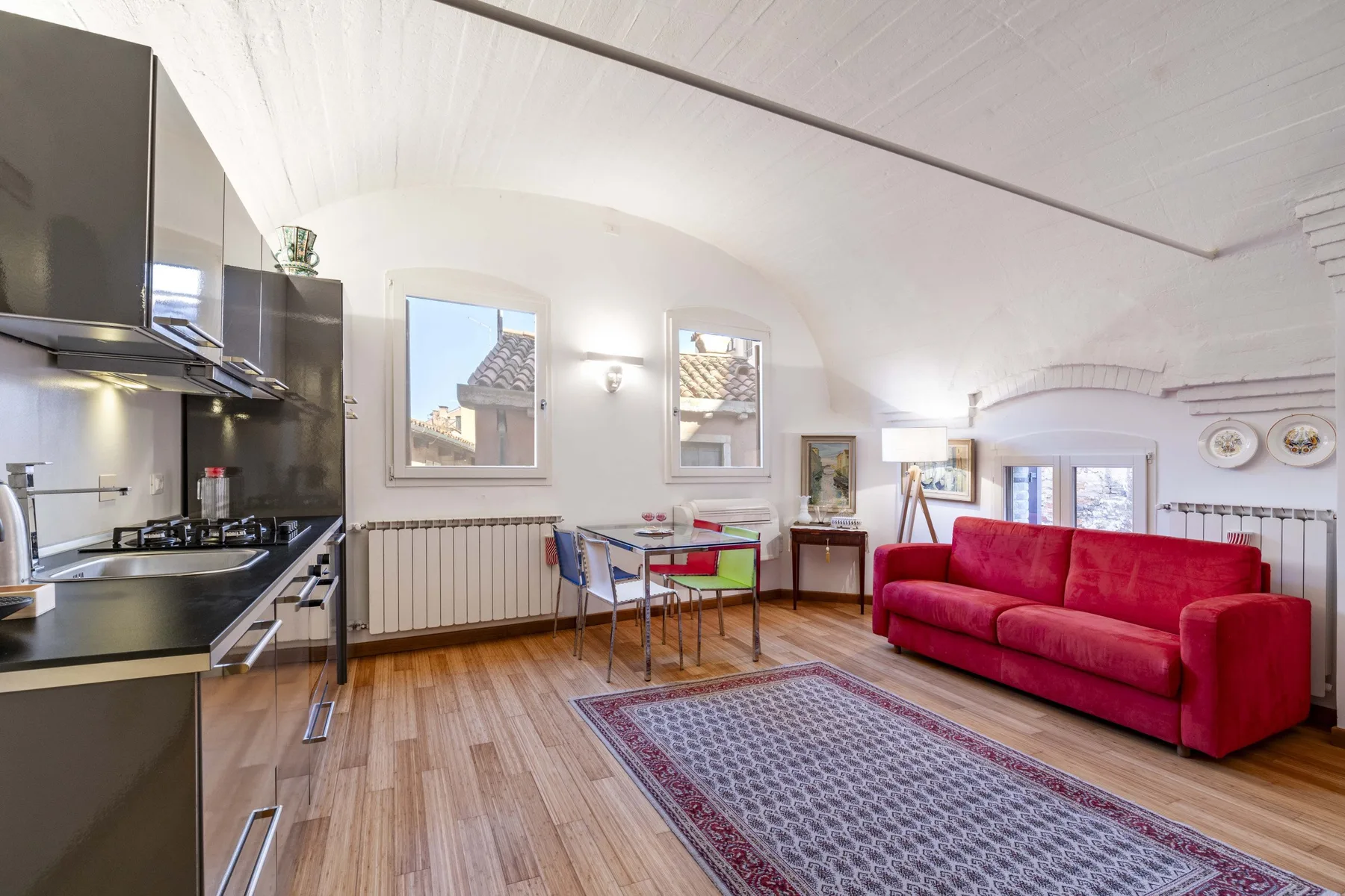 A bright open-plan living and dining area with a kitchen, red seating, and a rug.