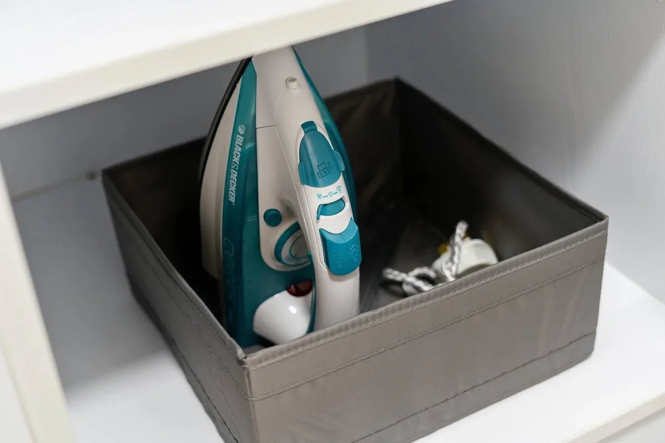 A storage bin beneath a shelf holds an iron and folded fabric.