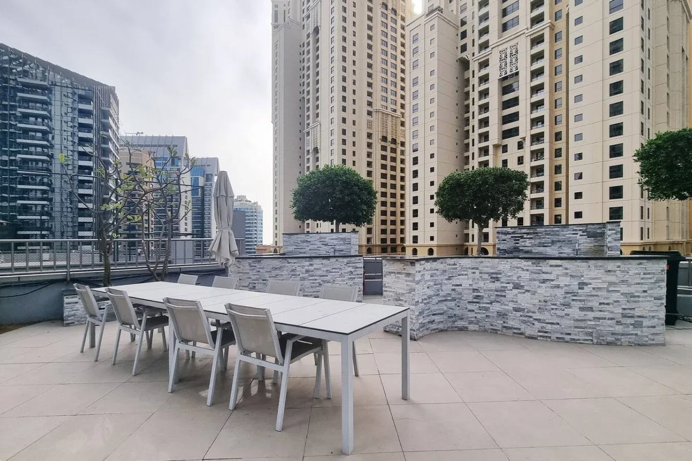 A rooftop terrace has an outdoor dining table beside planters and high-rise buildings.