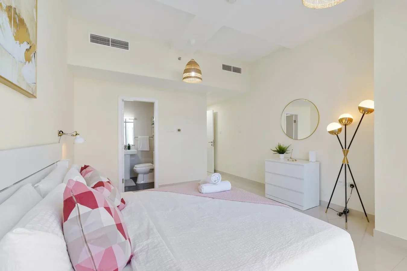 A bright bedroom with a bed, bedside lighting, and a dresser beneath a round mirror.