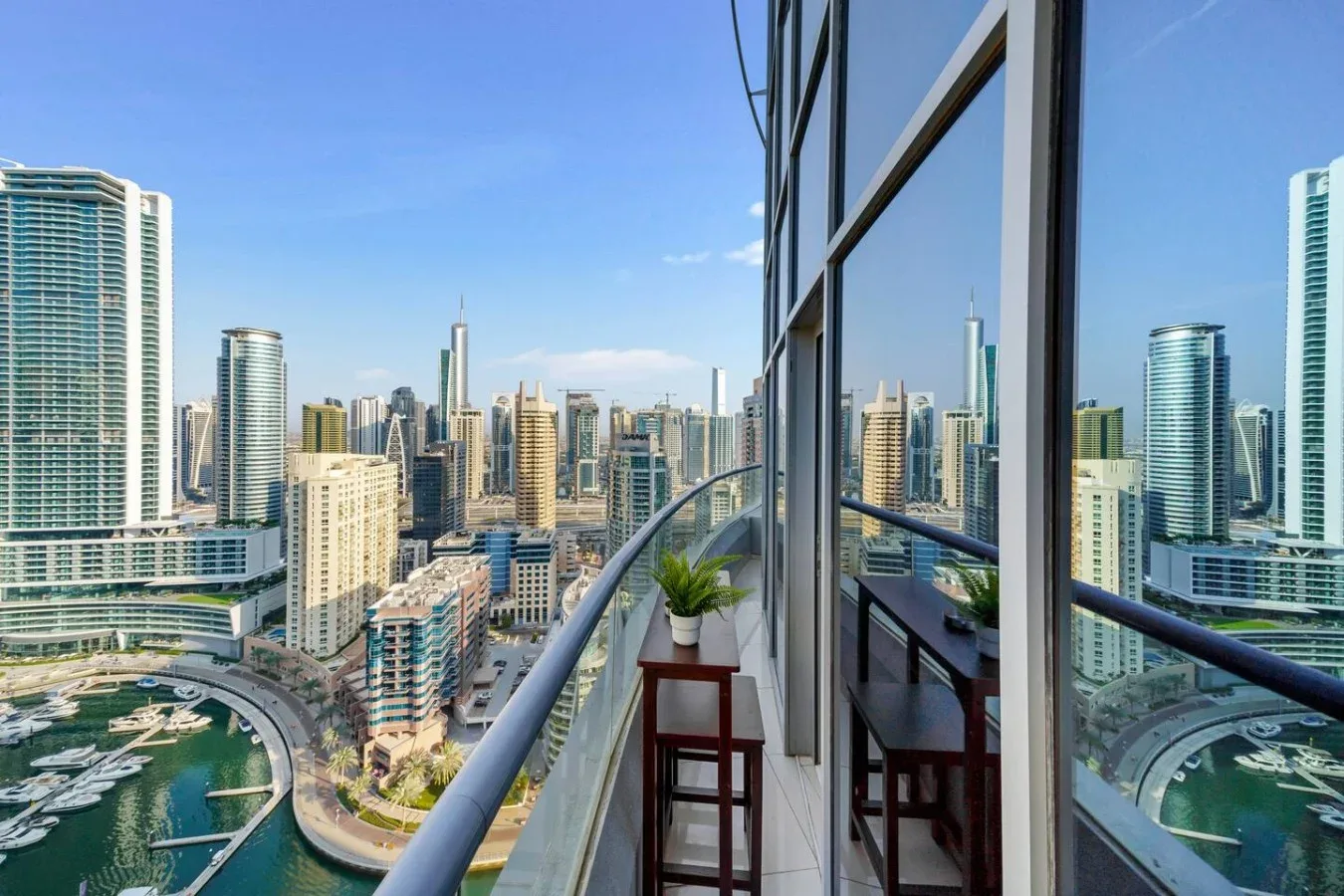 A high-rise balcony overlooks a dense city skyline, with a potted plant by the glass doors.