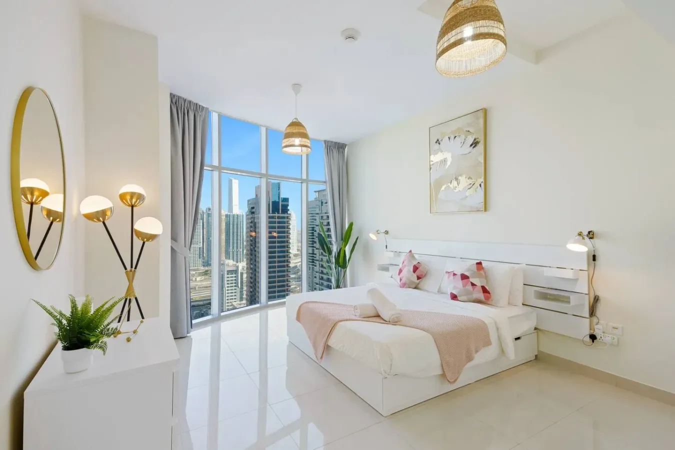 A bright bedroom with a bed, pale pink bedding, and large windows overlooking the city.
