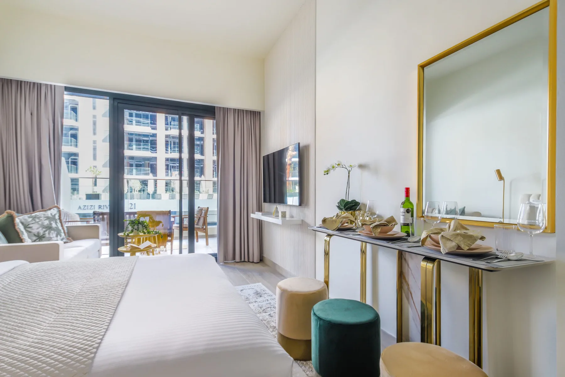 Un dormitorio luminoso con cama, asientos y una gran ventana frente a edificios cercanos.