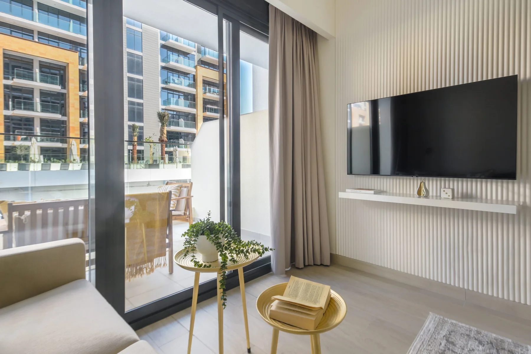 A bright living room with a wall-mounted television and floor-to-ceiling windows.