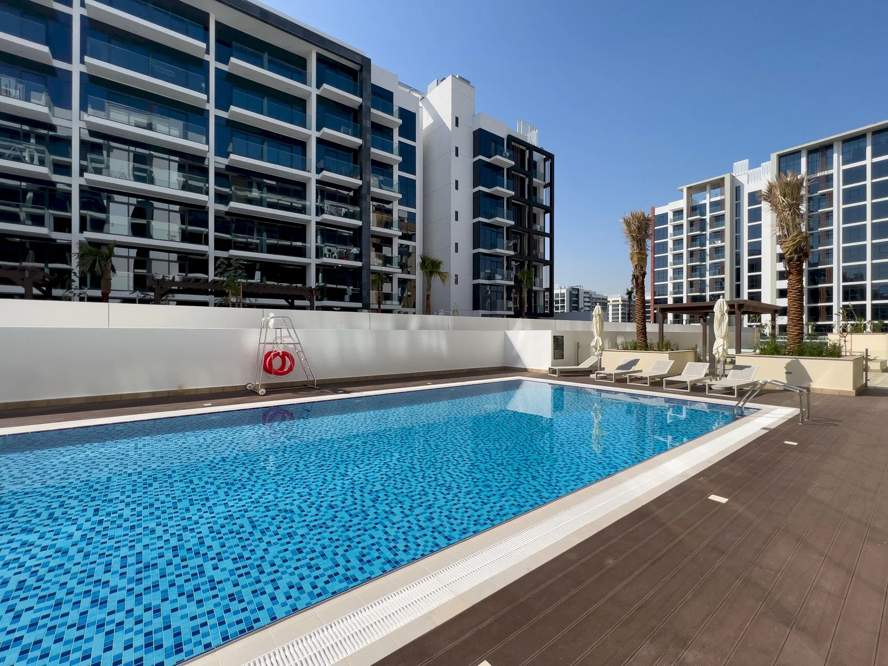 Una piscina al aire libre está rodeada por una terraza y edificios de apartamentos modernos.