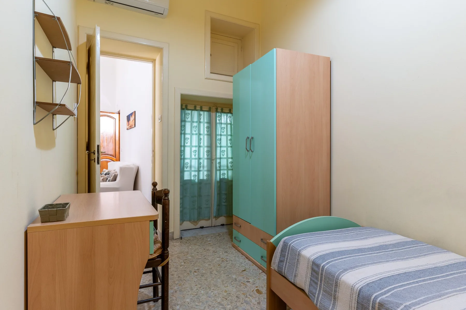 A compact bedroom with a single bed, desk, wardrobe, and curtained window.