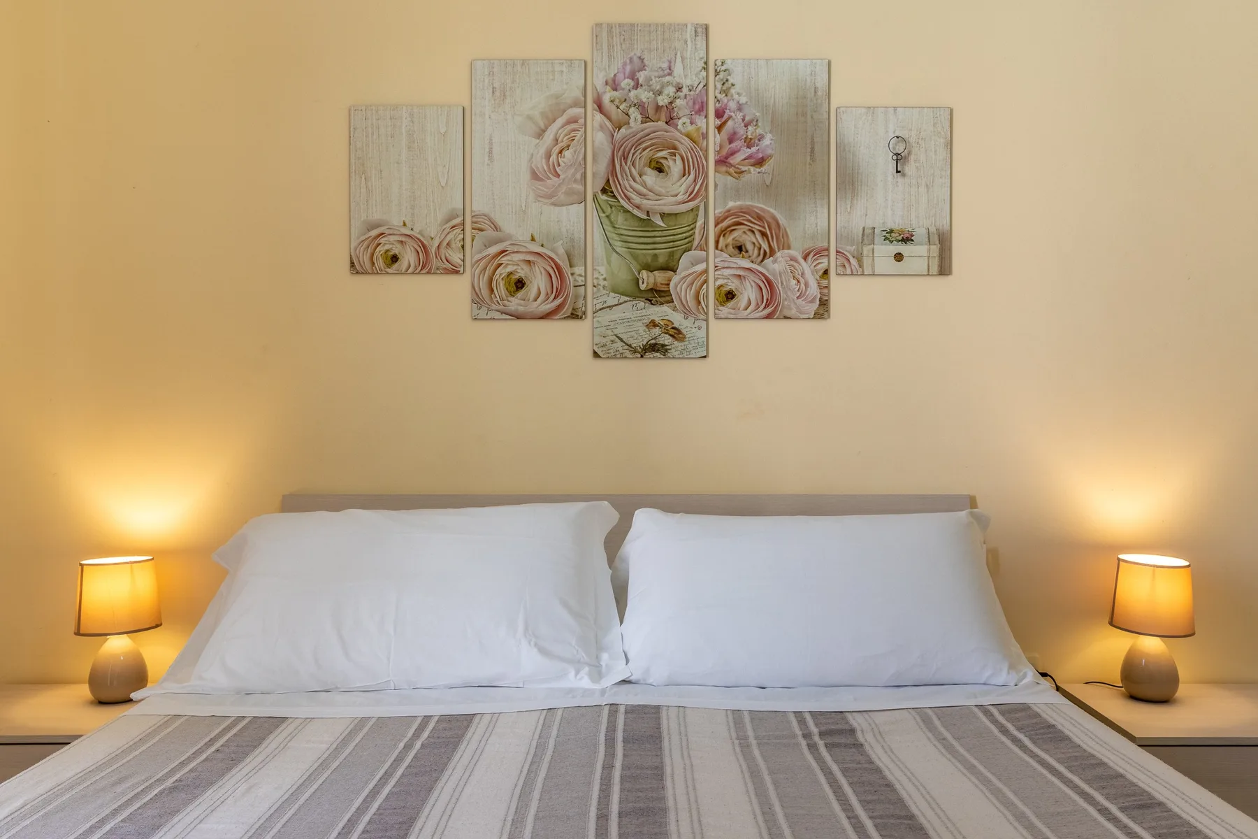 A bedroom with a neatly made bed, bedside lamps, and floral wall art.