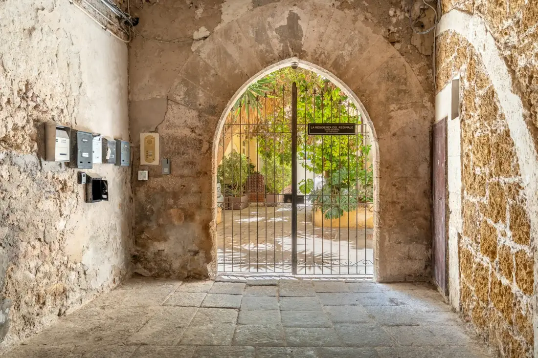 A stone passageway with an arched metal gate opening onto a leafy courtyard.