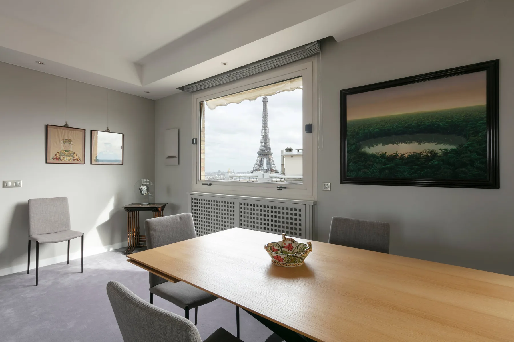 A dining area with a wooden table and chairs sits beside a window overlooking the Eiffel Tower.