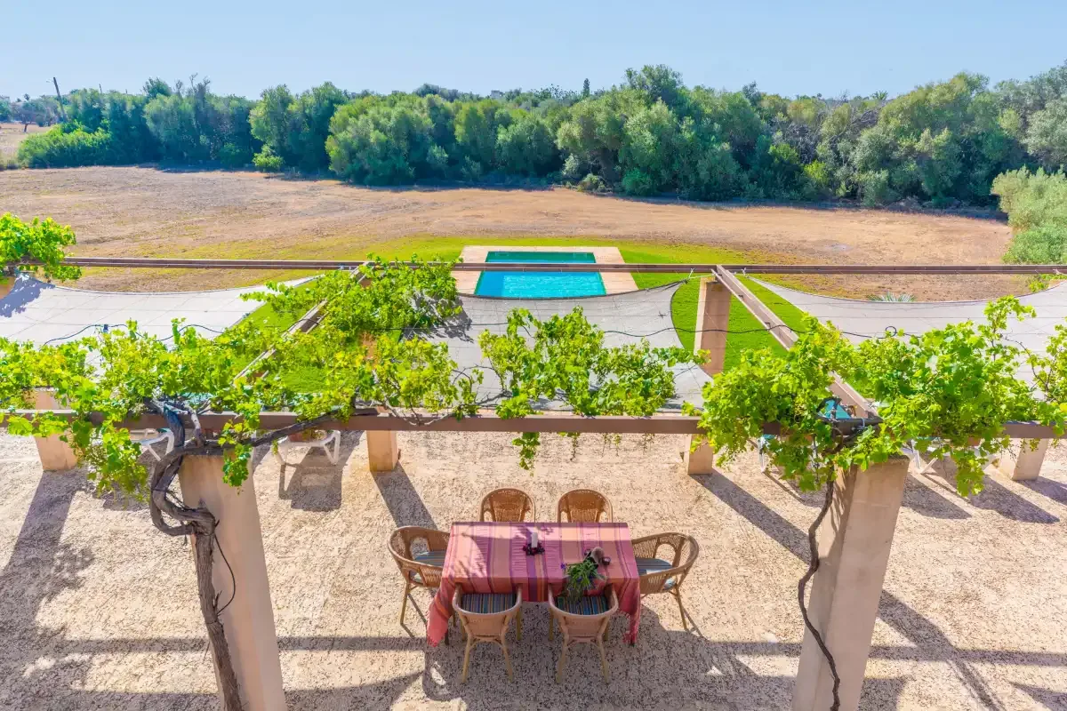An outdoor dining area beneath a vine-covered pergola overlooks a swimming pool and open landscape.