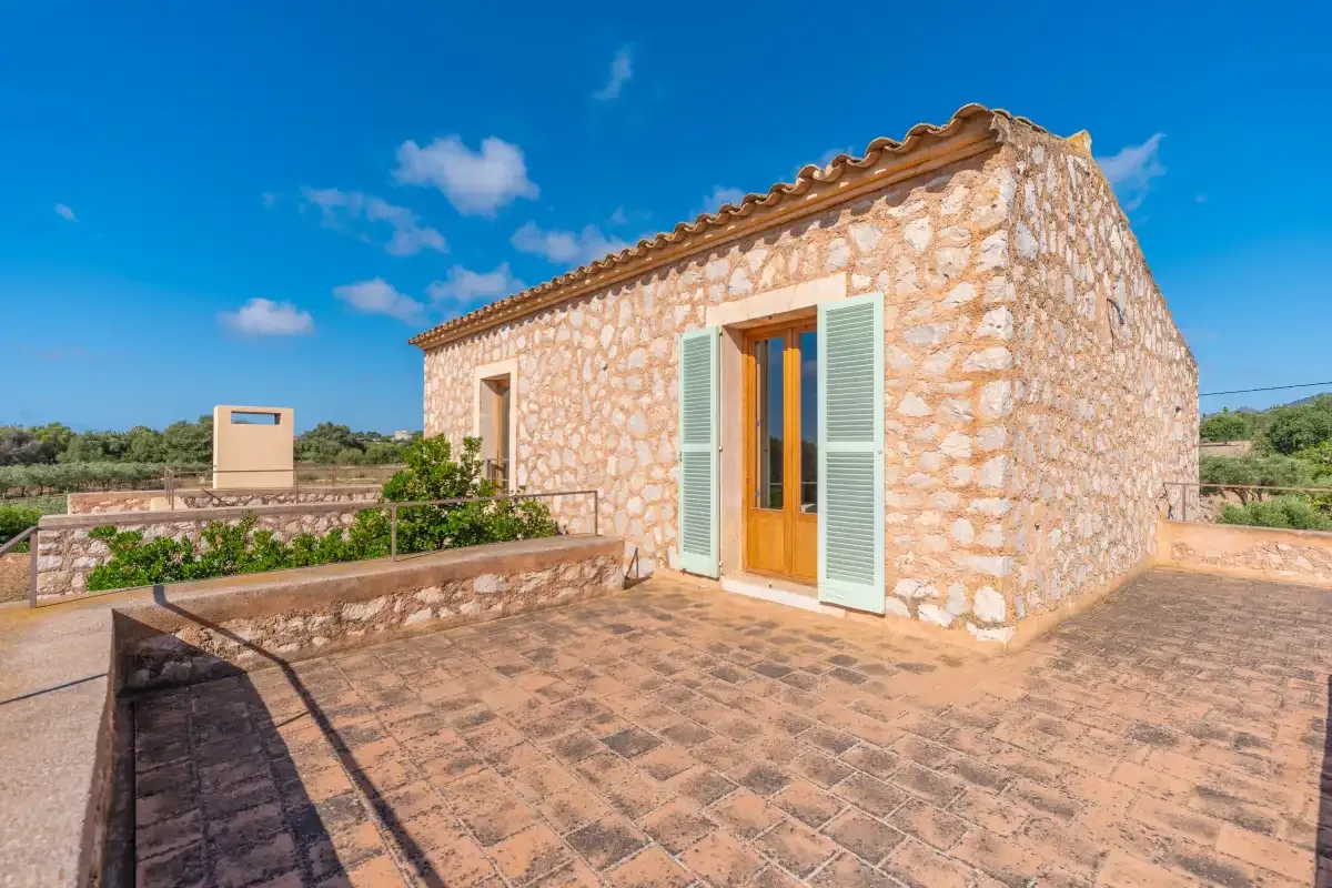 A stone building opens onto a sunlit paved terrace under a clear blue sky.