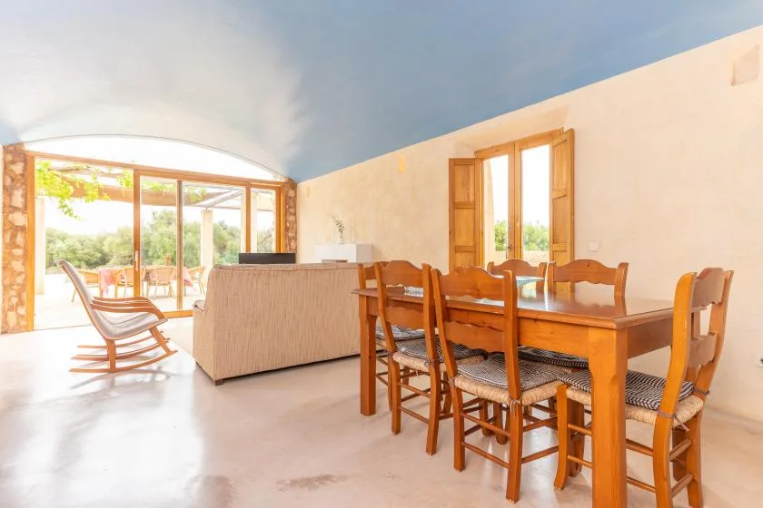 A bright living and dining area with wooden seating and large windows overlooking greenery.