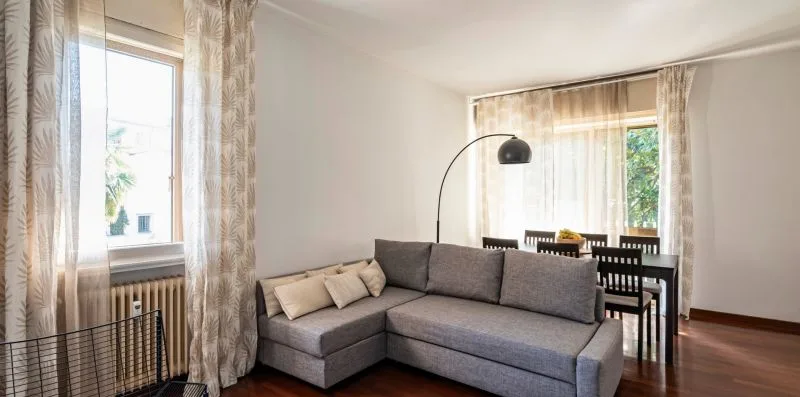 Una sala de estar con asientos seccionales grises y una mesa de comedor junto a las ventanas.