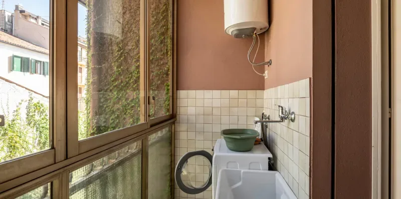 Un área de lavandería estrecha con una lavadora junto a ventanas altas.
