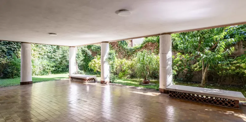 Un patio cubierto con suelo de baldosas, lados abiertos y vegetación de jardín.