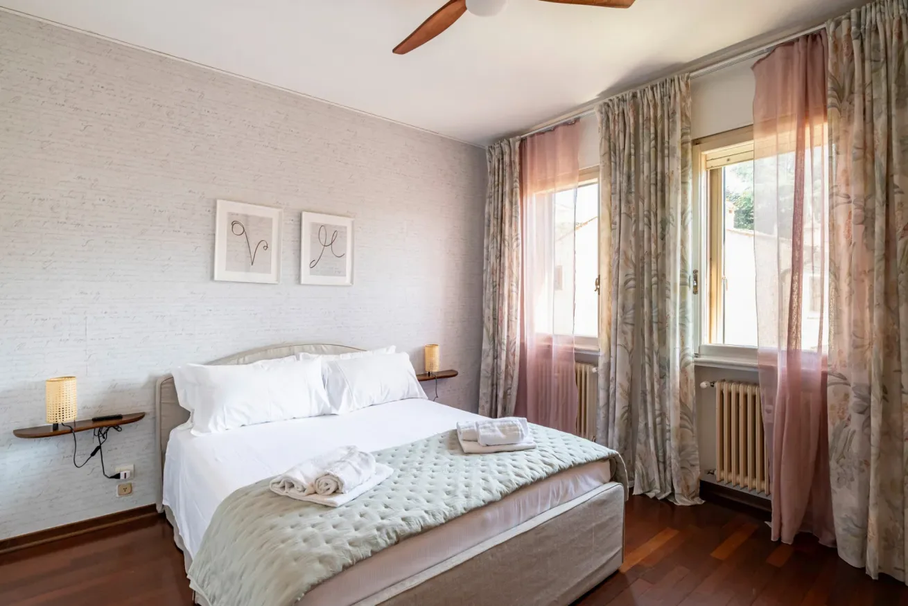 Un dormitorio con una cama vestida de blanco y cortinas junto a las ventanas.
