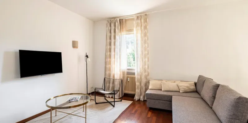 Una sala de estar con sofá seccional, televisor montado en la pared y ventana.