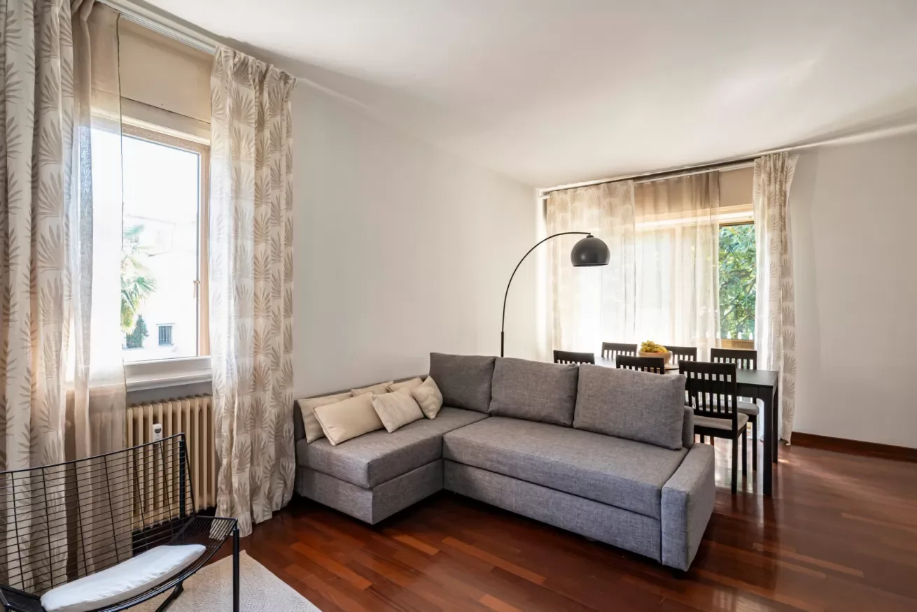 Una sala luminosa con un sofá seccional gris y una mesa de comedor junto a la ventana.