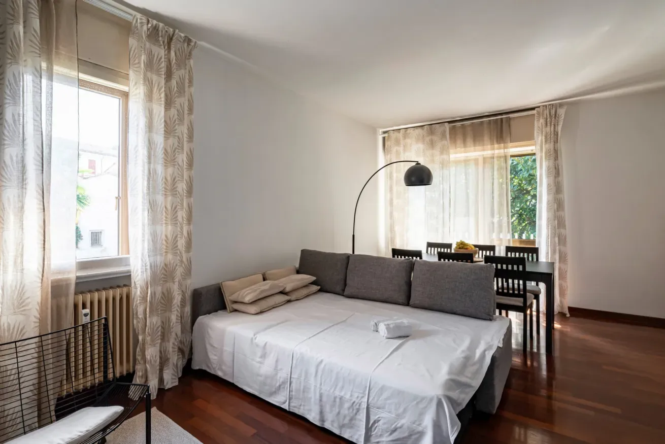 Una cama está junto a una mesa de comedor en una habitación luminosa con ventanas con cortinas.