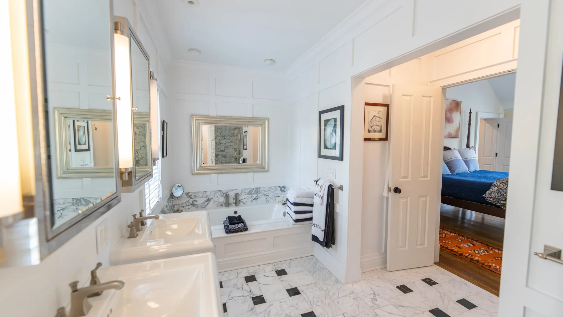Un baño luminoso con bañera, lavabos y una puerta hacia un dormitorio.