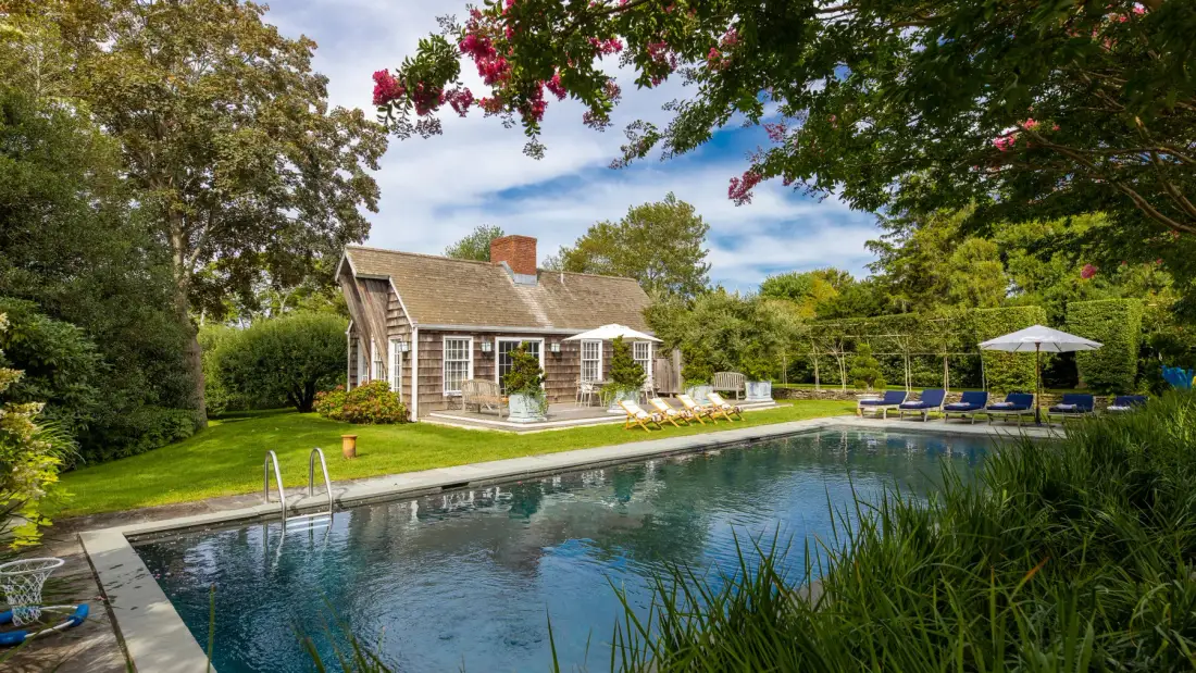 Una piscina junto a una casa pequeña, rodeada de árboles y ramas con flores.