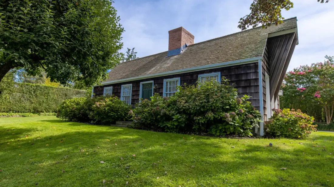 Una casa está junto a un césped verde, con arbustos a lo largo del exterior.