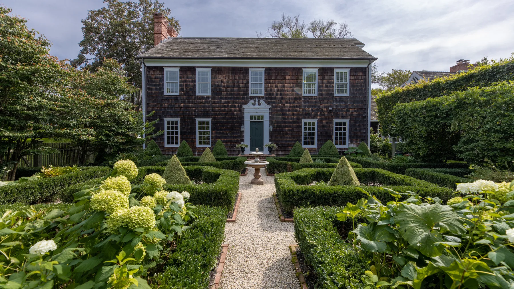Un sendero de grava atraviesa un jardín formal y conduce a una casa de tejas oscuras.