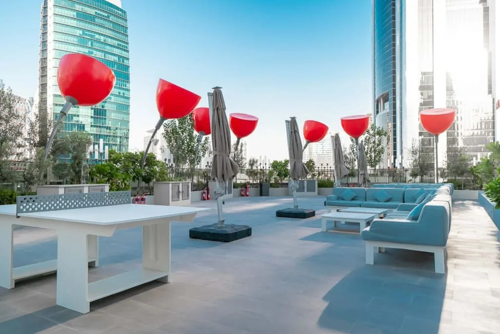 Una terraza al aire libre tiene asientos turquesa, sombrillas cerradas y lámparas escultóricas rojas.