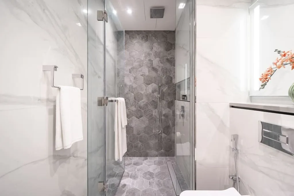 Una ducha de vidrio con azulejos grises estampados y paredes blancas con aspecto de mármol.