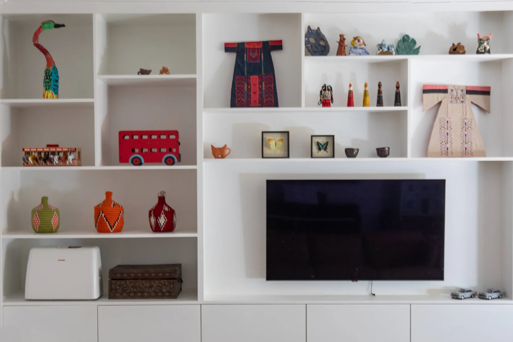A television is mounted beneath white display shelves holding colorful decorative objects.