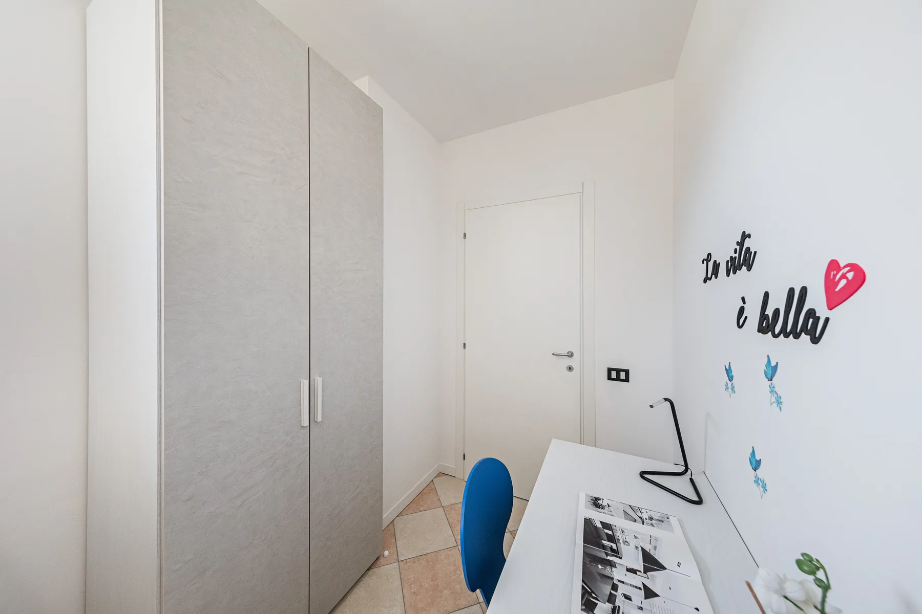 Un rincón luminoso de dormitorio con un armario alto, un escritorio y adornos en la pared.