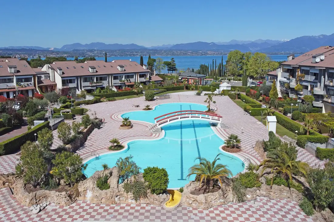 Una piscina exterior ajardinada está rodeada de edificios residenciales, con un lago y montañas al fondo.